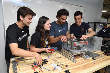 Four students working on their prototype device for Design Day. The device sits on top of a table; it is metal and has many wires attached to it.. 
