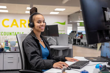 A Quantum health care coordinator is seated at a computer on her desk and is wearing a head set.ing a headset